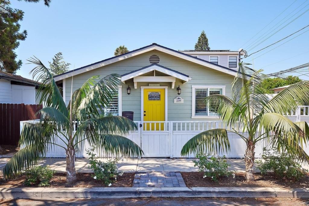 A seafoam-green cottage with white trim and a yellow front door, white fence, and two palm trees out front